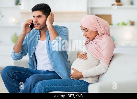 Worried Middle Eastern husband having phone call and supporting pregnant wife suffering prenatal contractions while sitting on sofa in living room ... Stock Photo