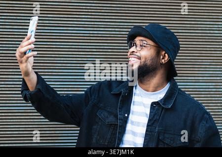 Black man with beard wearing eyeglasses and bucket hat taking selfie on smartphone standing in front of metal wall on street Stock Photo