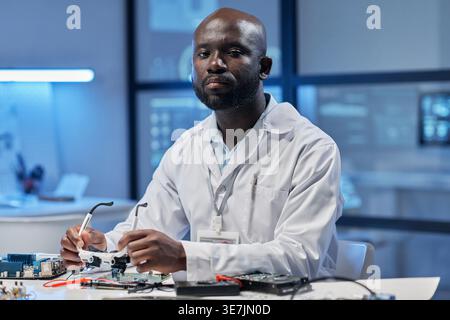 Black male computer engineer holding magnifying eyeglasses while sitting at table with computer components in laboratory Stock Photo