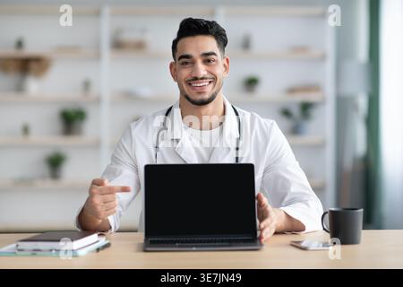 Smiling Middle Eastern male doctor wearing white robe with stethoscope showing and pointing at laptop sitting at table in clinic Stock Photo