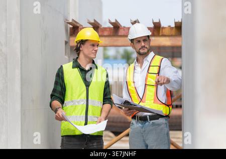 Hispanic male engineer with hardhat and safety vest discussing project with caucasian male colleague in construction site Stock Photo