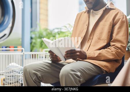 Cropped image Black man reading book while sitting on chair near washing machine in laundry shop Stock Photo