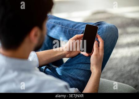 Arab man with beard using smarrtphone sitting with crossed legs on sofa in living room at home Stock Photo