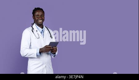 Smiling Black male doctor with dreads wearing white suit with stethoscope holding tablet while standing against purple background in studio Stock Photo
