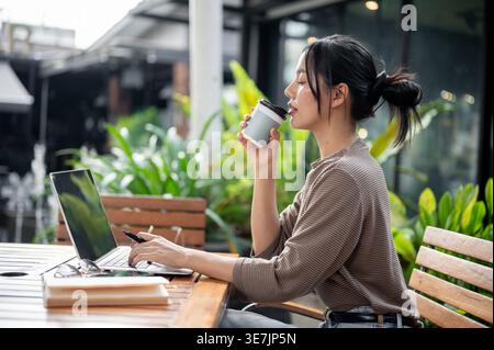 Asian woman drinking coffee while using laptop sitting at table in outdoor cafe Stock Photo