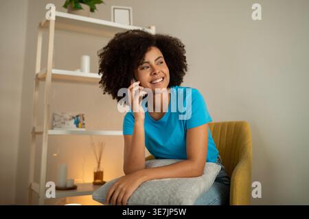 Black woman with curly afro having phone call and looking sideways sitting on chair with throw pillow against white wall at home Stock Photo