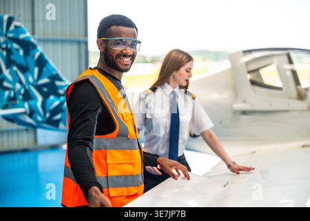 Smiling African American male aircraft engineer and female pilot inspecting airplane wing in hangar Stock Photo