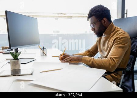 Black male architect with beard wearing brown turtleneck and eyeglasses working on blueprints for new project while sitting at desk with computer i... Stock Photo