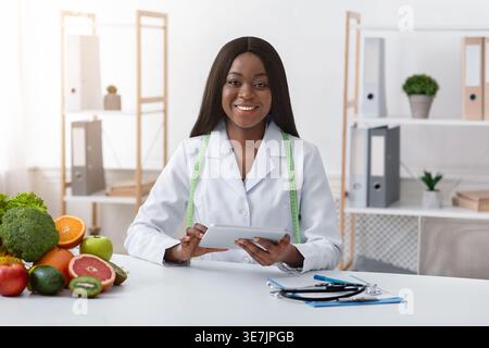 Black female nutritionist with tape measure around her neck holding tablet sitting at table with fruits and vegetables in clinic Stock Photo