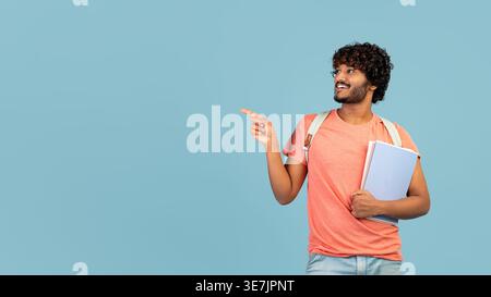 Bearded Indian male college student with curly hair pointing finger sideways holding books while standing in front of blue background in studio Stock Photo
