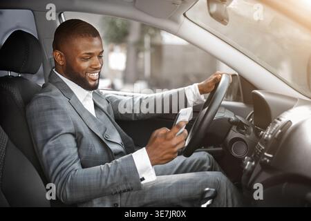 Smiling Black businessman using smLifestylephone sitting in car while driving during daytime Stock Photo