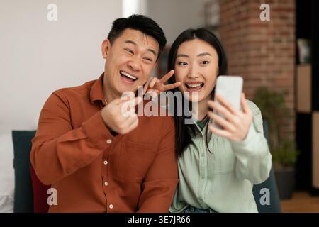 Asian couple gesturing peace signs having video call on smartphone while sitting together on sofa in living room at home Stock Photo