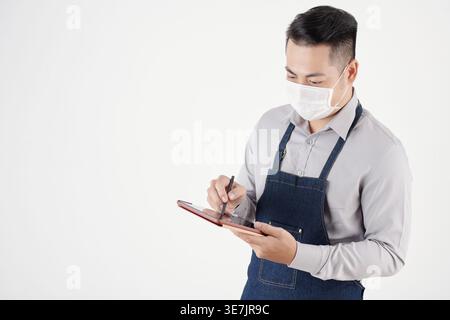 Asian male waiter wearing face mask and apron using tablet with stylus pen standing against white background Stock Photo