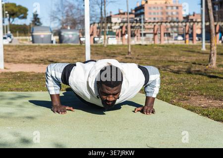 Mixed-race man with beard wearing jacket doing push-ups in park on sunny day Stock Photo