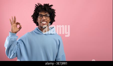 Smiling Black man with afro wearing blue hoodie and eyeglasses standing against pink wall in studio Stock Photo