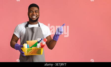 Black man with beard wearing apron and rubber gloves carrying basket of cleaning supplies pointing away standing against pink background in studio Stock Photo