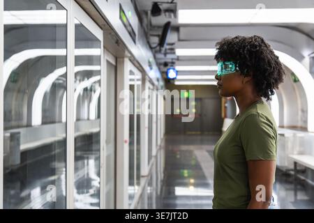 Black woman with afro wearing futuristic smart glasses and looking away while standing sideways in subway station Stock Photo