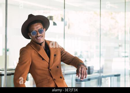 Hispanic man wearing brown suit, sunglasses, and fedora hat standing and leaning on glass wall outside the building in city during daytime Stock Photo