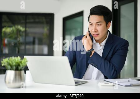 Asian businessman having phone conversation while working on laptop sitting at table in office during daytime Stock Photo