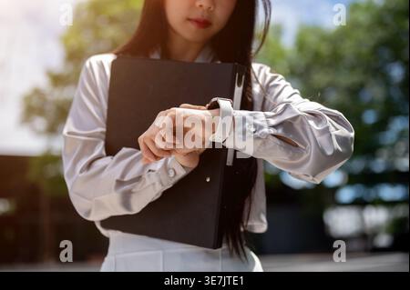 Thai business woman holding clipboard near chest looking at watch late for work standing on street in city during daytime Stock Photo