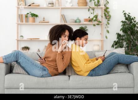 Black woman with shocked expression holding smartphone looking at smartphone using by female friend using smartphone while sitting back to back on ... Stock Photo