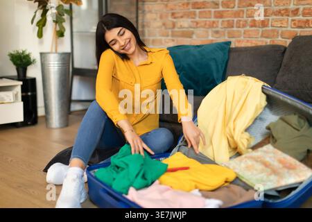 Smiling Middle Eastern woman having phone call while folding clothes in suitcase sitting on floor near sofa in living room at home Stock Photo