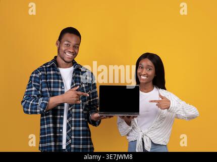 Black couple holding and pointing fingers at laptop while standing against yellow background in studio Stock Photo