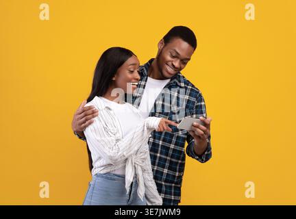 Black couple using smartphone together while standing against yellow background in studio Stock Photo