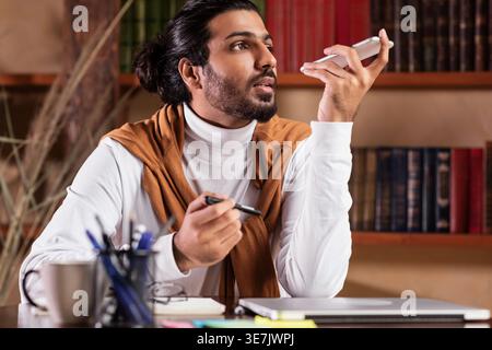 Indian male teacher recording voice message using smartphone sitting at table in library Stock Photo