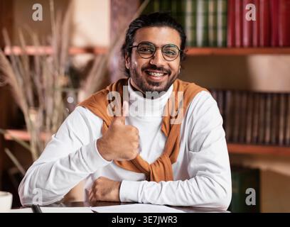 Indian male student wearing eyeglasses showing thumbs up sitting at table in library Stock Photo