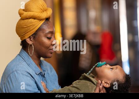 Black mother wearing yellow head wrap and long sleeve denim shirt holding and playing with her baby boy with pacifier while sitting at home Stock Photo