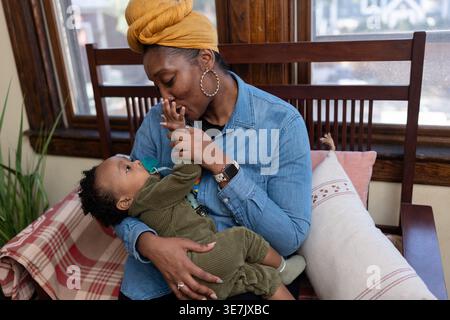 Black mother wearing yellow head wrap and long sleeve denim shirt carrying and playing with her baby boy with pacifier while sitting on wooden sofa... Stock Photo