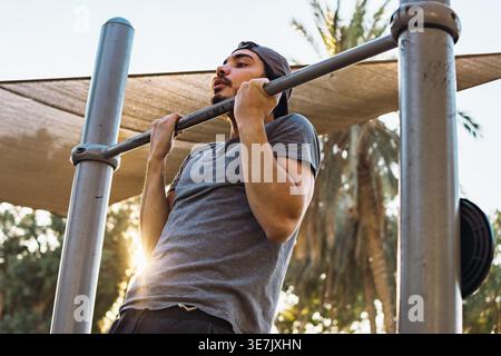 Middle Eastern man with beard wearing cap doing pull-ups in outdoor gym during sunny day Stock Photo