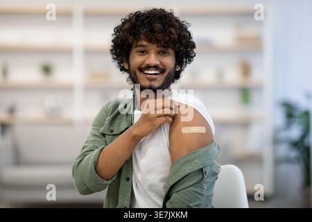 Asian man with curly hair showing bandage on her arm getting vaccinated while sitting on chair in clinic Stock Photo
