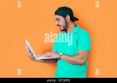 Arab man with beard wearing green cap and shirt using tablet with keyboard while standing against orange background in studio Stock Photo