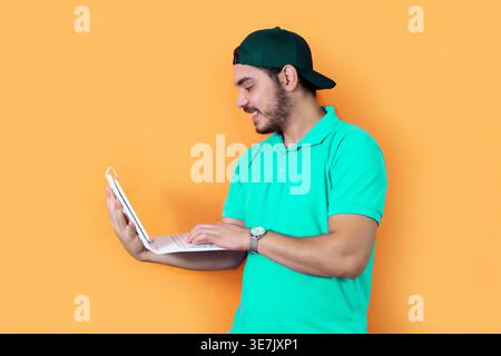 Arab man with beard wearing green cap and shirt using tablet with keyboard while standing against yellow background in studio Stock Photo