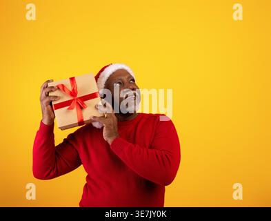 Middle-aged Black man wearing Santa hat looking up and shaking gift box near his ear while standing against yellow background in studio Stock Photo