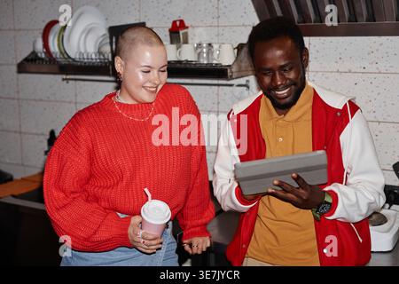 Black male college student using tablet leaning on kitchen counter next to caucasian female friend holding reusable cup while standing together in ... Stock Photo