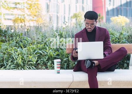 Hispanic businessman wearing maroon suit and eyeglasses using smartphone and laptop while sitting with crossed legs on concrete bench in city park ... Stock Photo