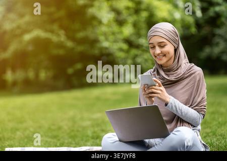 Arab woman wearing hijab using smartphone while sitting with laptop on picnic blanket in park during daytime Stock Photo