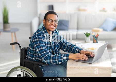 Smiling Black disabled man wearing eyeglasses working on laptop while sitting at table in home office Stock Photo