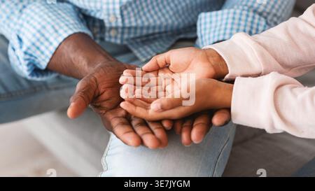 Cropped image of Black granddaughter with palms open on grandfather's hands placed on his lap while sitting on sofa at home Stock Photo