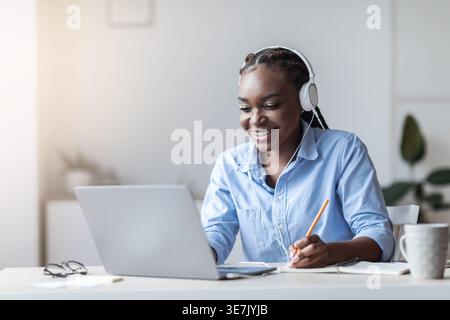 Black business woman with braids wearing headphones using laptop taking notes while sitting at table in home office Stock Photo