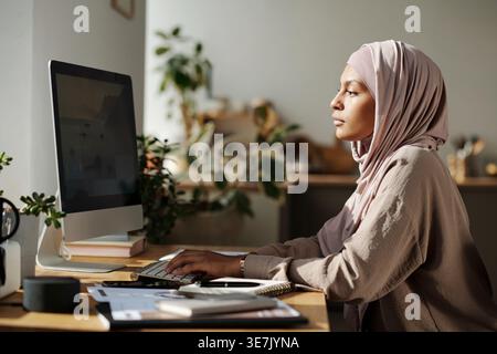 Black female programmer in hijab working on computer while sitting at table with smLifestylephone and notepad at home office Stock Photo