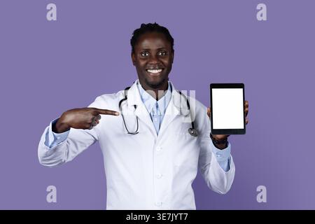 Smiling Black male doctor with dreads wearing white suit with stethoscope pointing at tablet on hand while standing against purple background in st... Stock Photo
