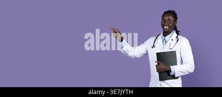 Smiling Black male doctor with dreads wearing white suit with stethoscope holding clipboard pointing away while standing against purple background ... Stock Photo