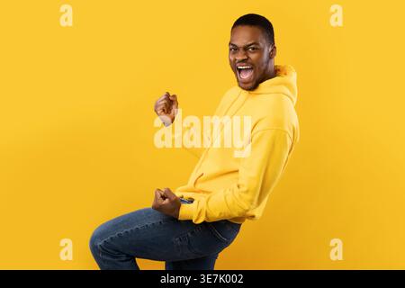 Emotional Black Man Shaking Fists In Joy Posing Over Yellow Background. Excited Guy Celebrating Success Shouting Gesturing Yes In Studio. Joy Of Victo Stock Photo
