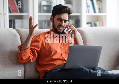 Upset Middle Eastern man with laptop gesturing hand talking on smartphone sitting on couch at home Stock Photo