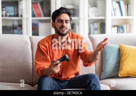 Disappointed Middle Eastern man gesturing hand playing video game sitting on sofa at home Stock Photo