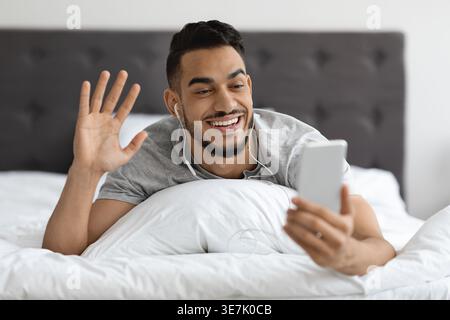 Arab man in earphones having video call using smartphone while lying on bed in bedroom at home Stock Photo